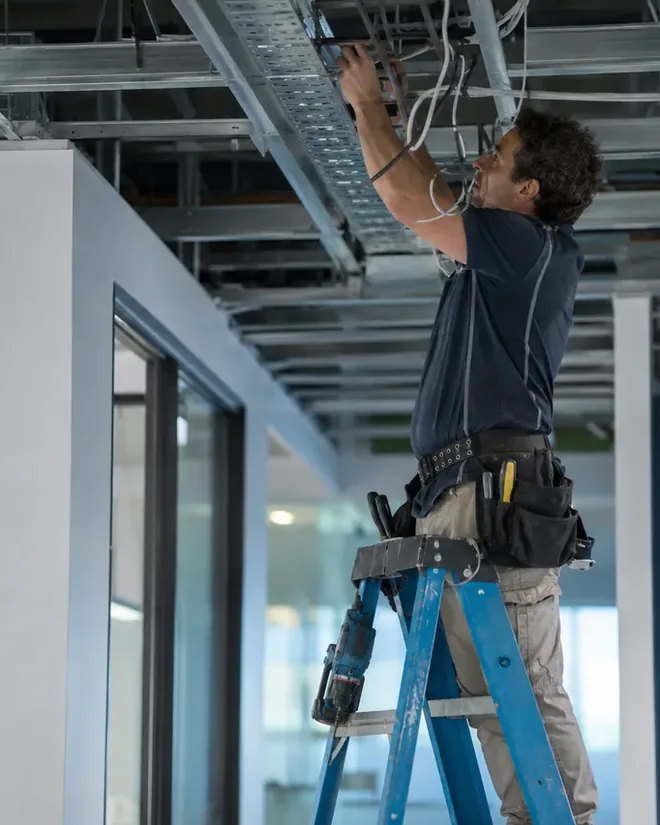 An electrician on a step ladder working on cabling in an exposed ceiling
