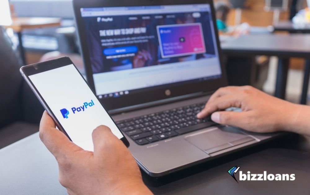 Hand of a businessman paying with smartphone and laptop displaying PayPal logo on screen